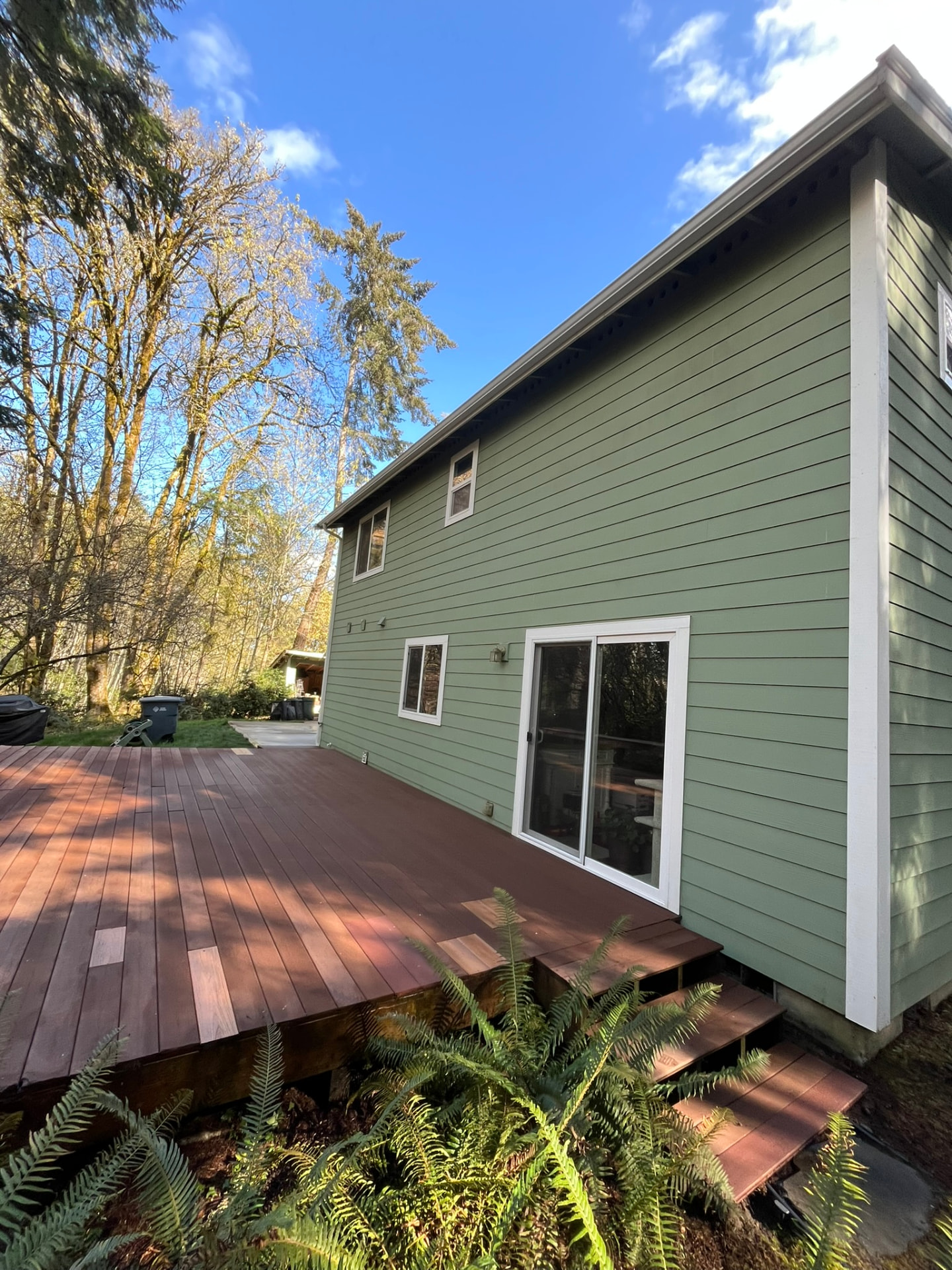 A clean sage-green two-storey house with bright white trim and a fresh wooden deck among tall trees