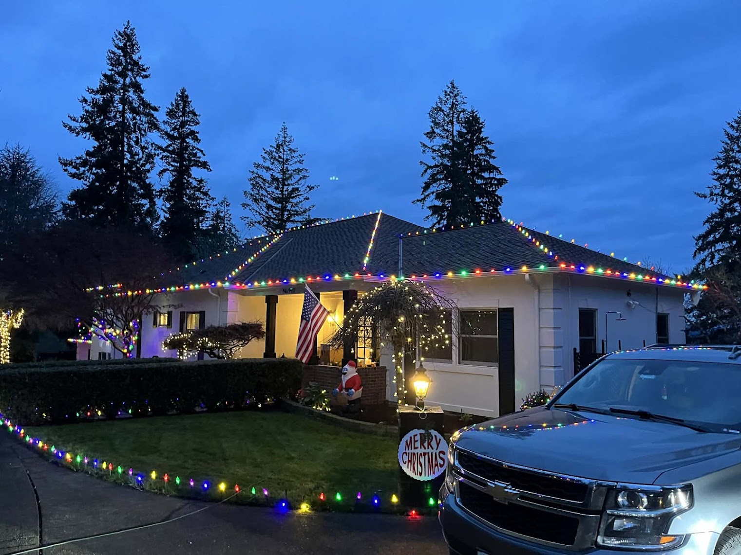 A white house at dusk with multicoloured lights along the roofline, hedge and driveway