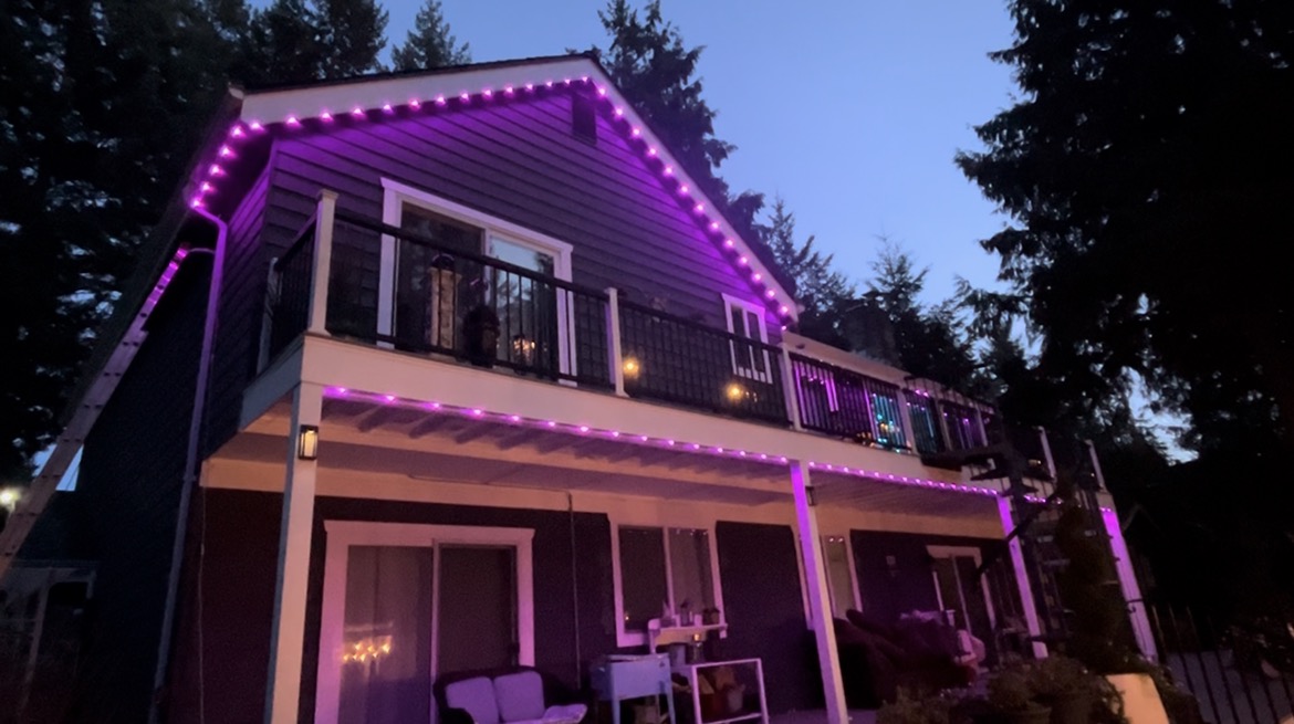 Purple permanent lights along the deck and roofline of a house at dusk